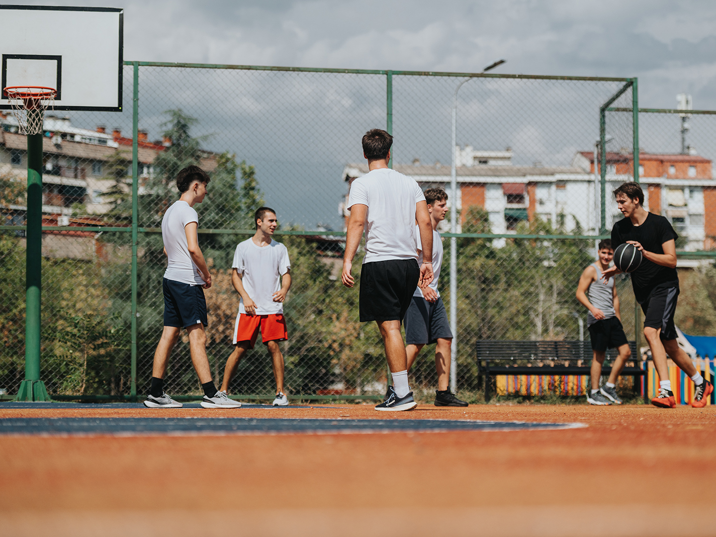 Sechs junge Männer spielen Basketball auf einem Außenplatz. Einige stehen in einer Gruppe, während ein Spieler den Ball dribbelt. Im Hintergrund sind Wohngebäude und Bäume sichtbar. Der Himmel ist bewölkt.