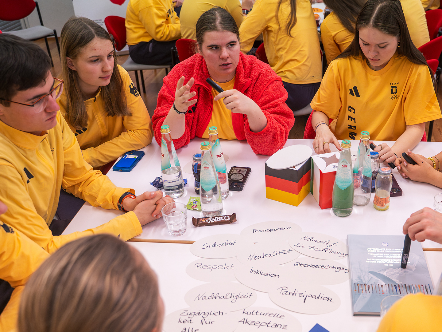 Eine Gruppe junger Menschen in gelben T-Shirts sitzt an einem Tisch. Sie diskutieren und arbeiten an einem Projekt. Auf dem Tisch liegen Wasserflaschen, Notizen und verschiedene Materialien. Einige Teilnehmer sind aktiv am Sprechen, während andere aufmerksam zuhören.
