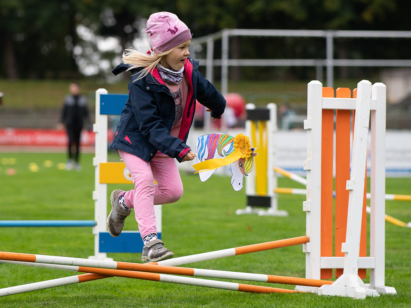 Ein Mädchen springt über ein Hindernis im Agility-Sport. Sie trägt eine Mütze und einen blauen Mantel, während sie ein Spielzeug in der Hand hält. Der Hintergrund zeigt eine Sportanlage mit weiteren Hindernissen und Zuschauern.