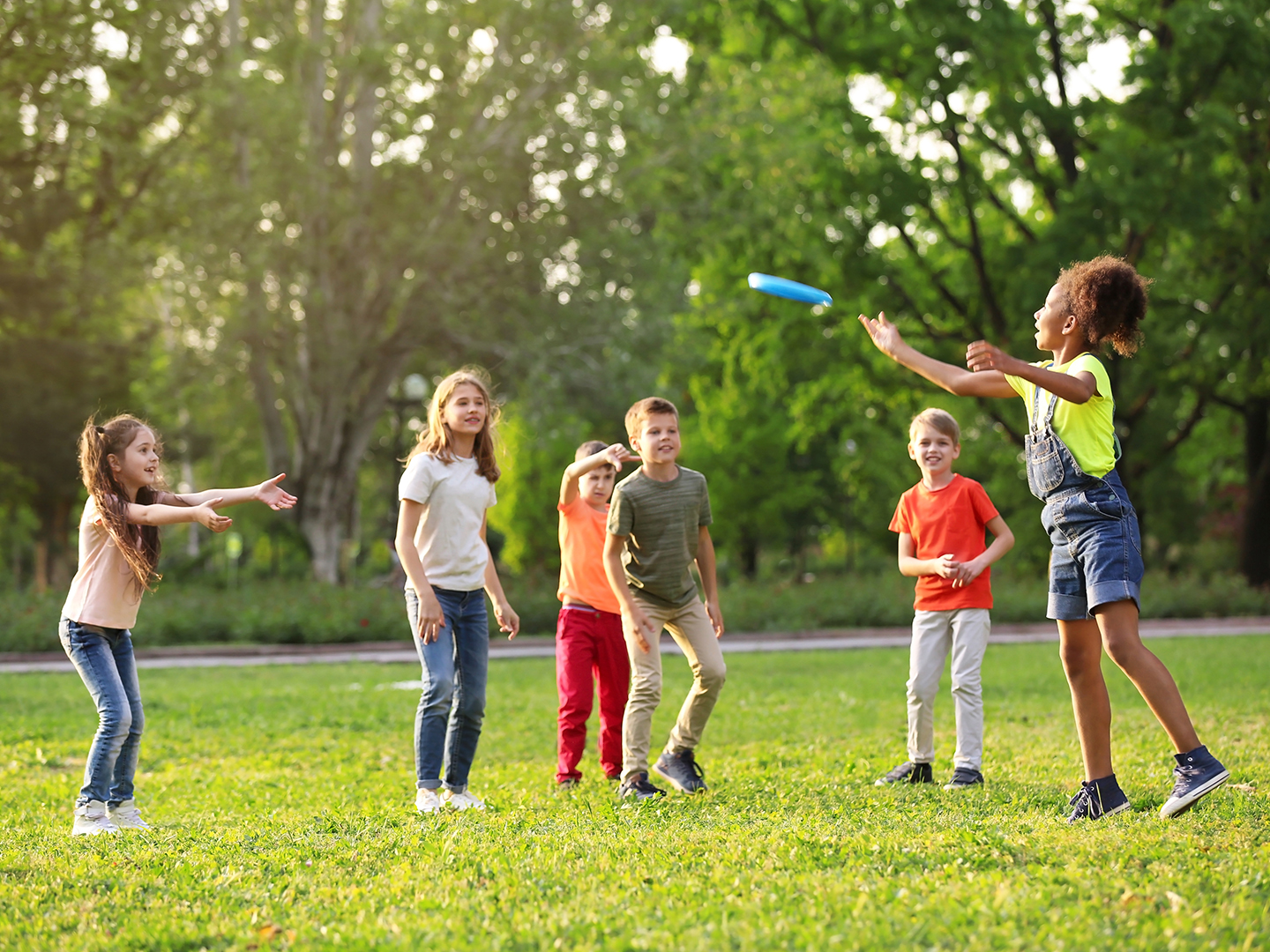 Sechs Kinder spielen im Freien Frisbee. Eine Mädchen wirft den Frisbee, während die anderen gespannt zuschauen. Die Szene findet auf einer grünen Wiese mit Bäumen im Hintergrund statt. Es ist ein sonniger Tag.