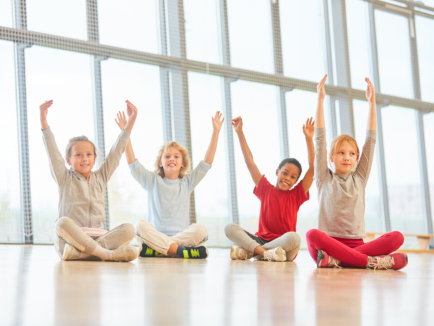 Vier Kinder sitzen auf dem Boden in einem hellen Raum mit großen Fenstern. Sie haben die Arme erhoben und lächeln. Die Kinder tragen bequeme Kleidung in verschiedenen Farben und scheinen Freude zu haben.