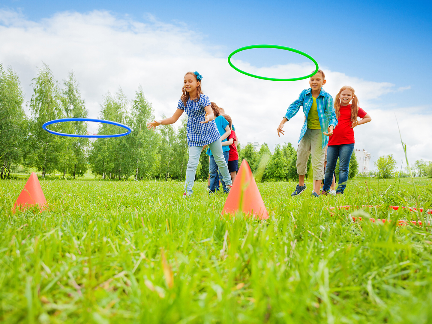 Eine Gruppe von Kindern spielt im Freien auf einer Wiese. Einige Kinder werfen Hula-Hoops, während andere zuschauen. Im Hintergrund sind Bäume und ein blauer Himmel zu sehen. Die Kinder tragen bunte Kleidung und wirken fröhlich.