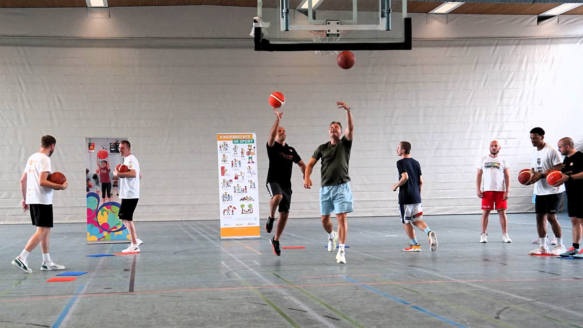 Gruppe von acht Personen in Sportkleidung, die in einer Halle Basketball spielen. Zwei Personen werfen einen Ball, während andere warten. Im Hintergrund ein Informationsbanner zur Fortbildung.