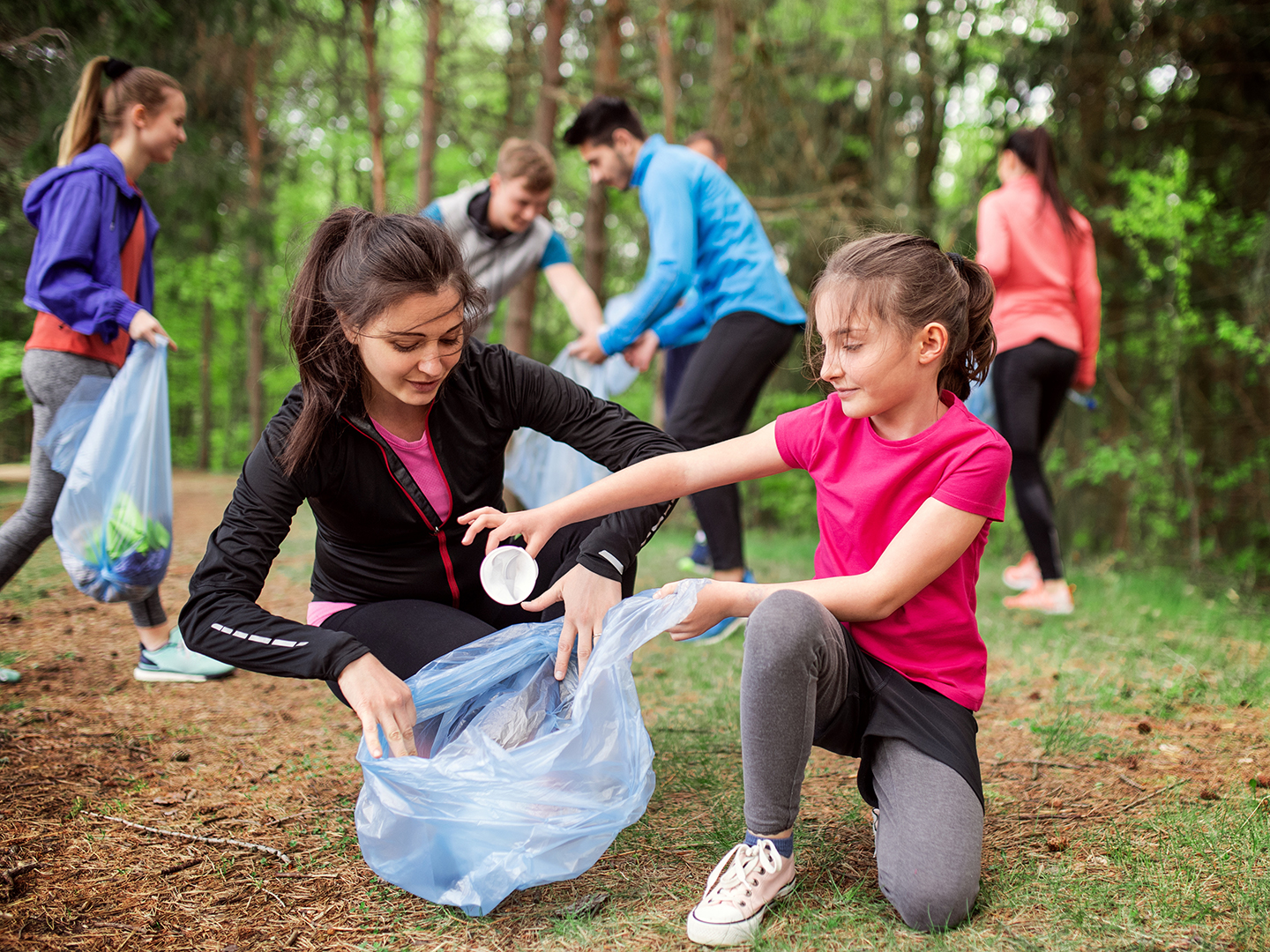 Eine Gruppe von Menschen sammelt Müll im Wald. Zwei Mädchen, eines in schwarzer und das andere in pinker Sportkleidung, arbeiten zusammen, um Abfall in einen blauen Müllsack zu legen. Im Hintergrund sind weitere Personen zu sehen, die ebenfalls Müll einsammeln.