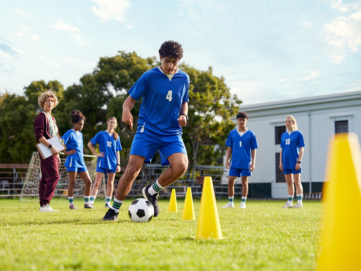 Ein junger Fußballspieler in blauer Sportkleidung dribbelt einen Ball um gelbe Hütchen auf einem Rasenplatz. Im Hintergrund stehen mehrere Teamkollegen und eine Trainerin, die das Training beobachten.