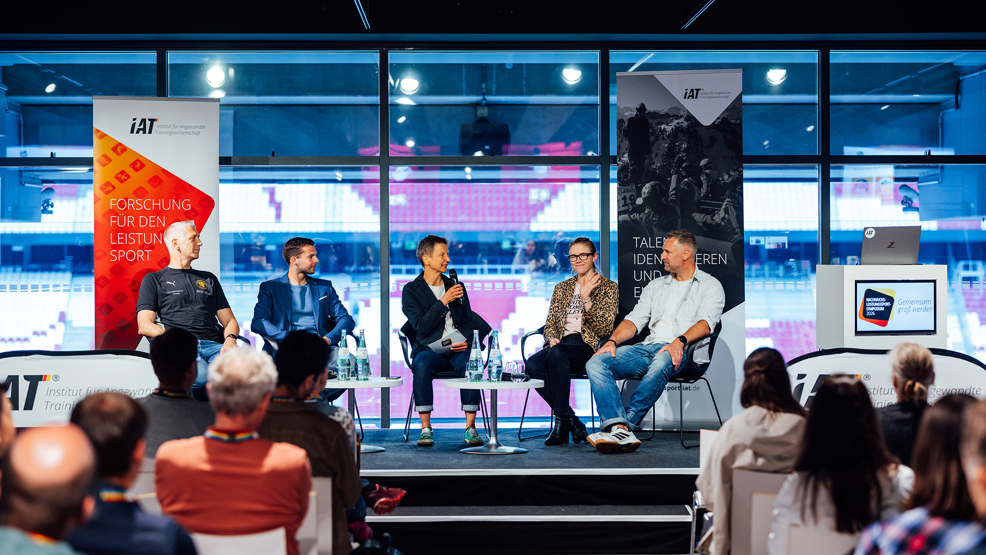 Podiumsdiskussion beim IAT NWLS Symposium: Fünf Personen sitzen auf einer Bühne vor Publikum und sprechen in Mikrofone. Im Hintergrund sind große Fenster sowie Banner mit IAT-Logo und Texten zur Sportforschung sichtbar.