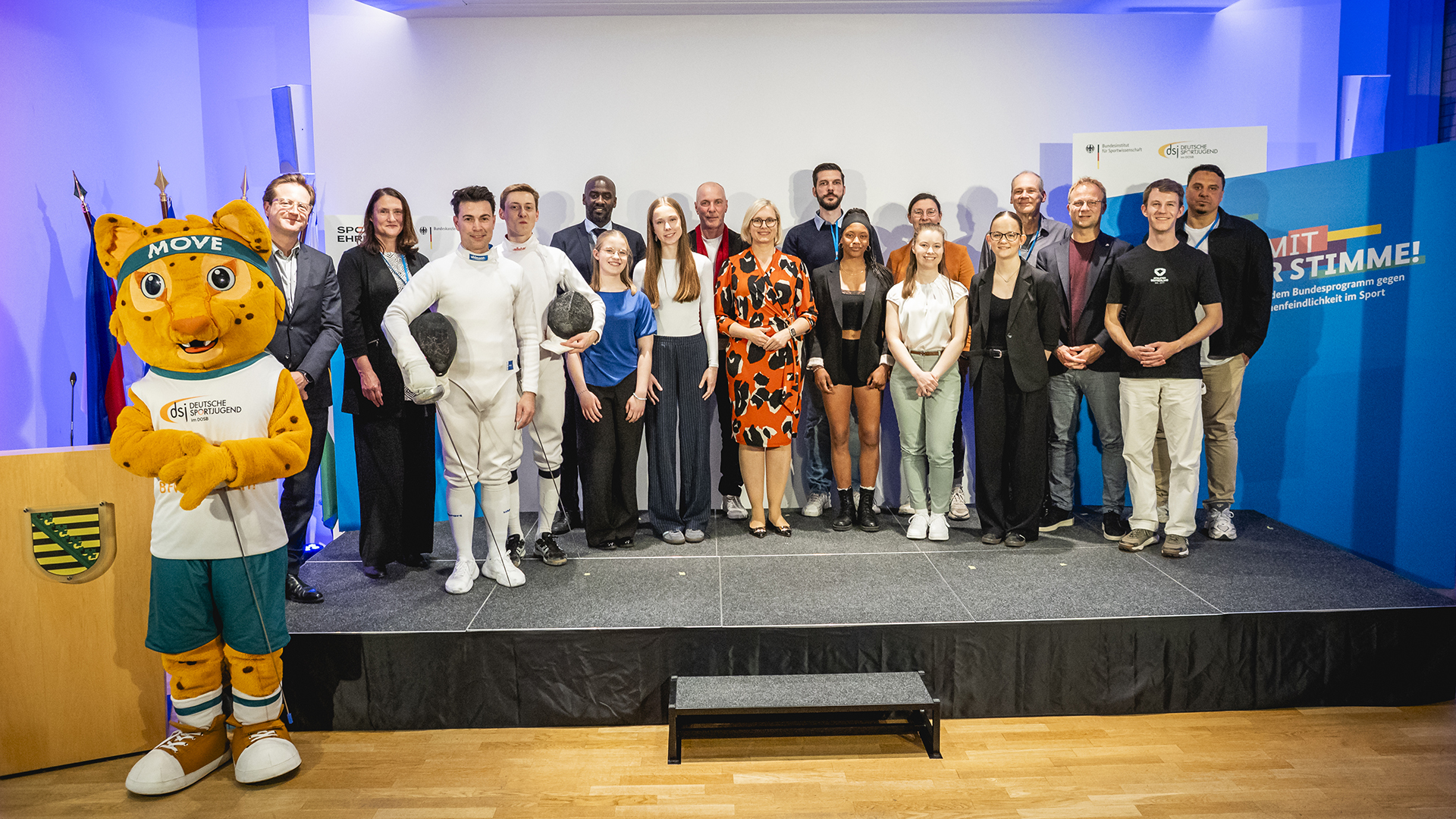 Gruppenfoto auf einer Bühne bei der Preisverleihung des Vereinspreises ‚Sport mit Haltung‘ 2026: Mehrere Personen stehen nebeneinander, darunter zwei Sportler in Fechtkleidung sowie ein Maskottchen; im Hintergrund sind Banner zum Bundesprogramm gegen Extremismus und Menschenfeindlichkeit im Sport zu sehen.