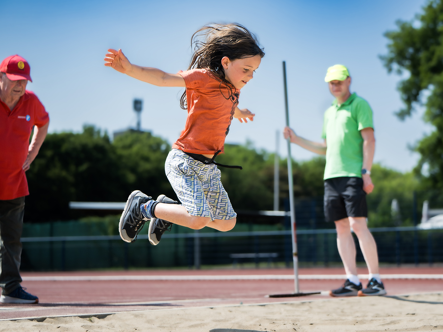 Ein Kind springt mit Freude in einen Sandgrube beim Weitsprung. Im Hintergrund stehen zwei Erwachsene, die den Sprung beobachten. Der Himmel ist blau und die Umgebung ist eine Sportanlage.