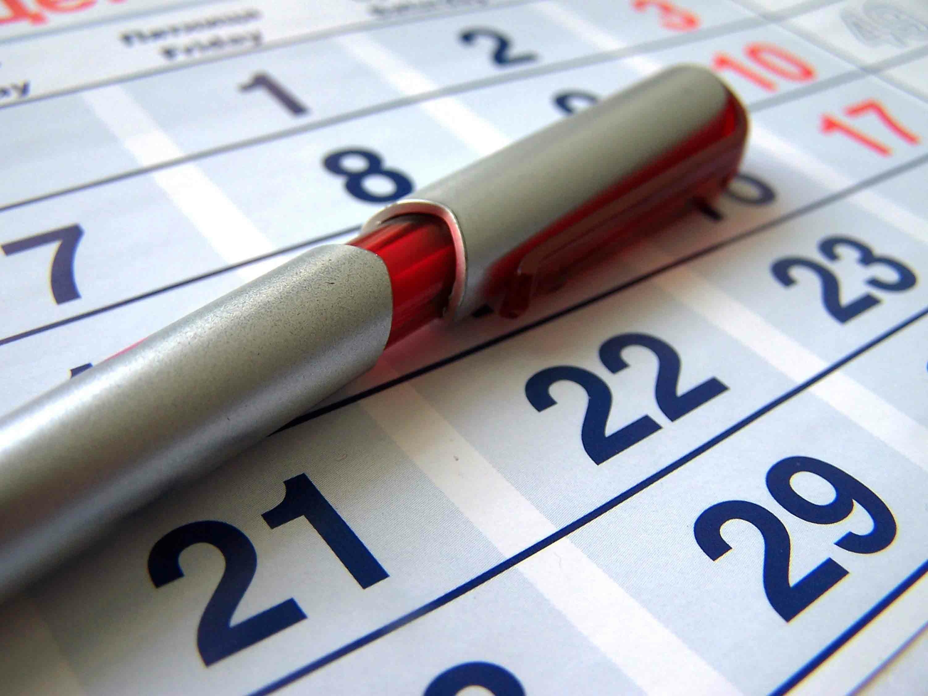 Ein silberner Kugelschreiber liegt auf einem Kalender, der die Tage 21 bis 29 anzeigt. Die Zahlen sind in blauer Schrift auf einem weißen Hintergrund gedruckt. Der Kalender zeigt den Monat und die Wochentage an.