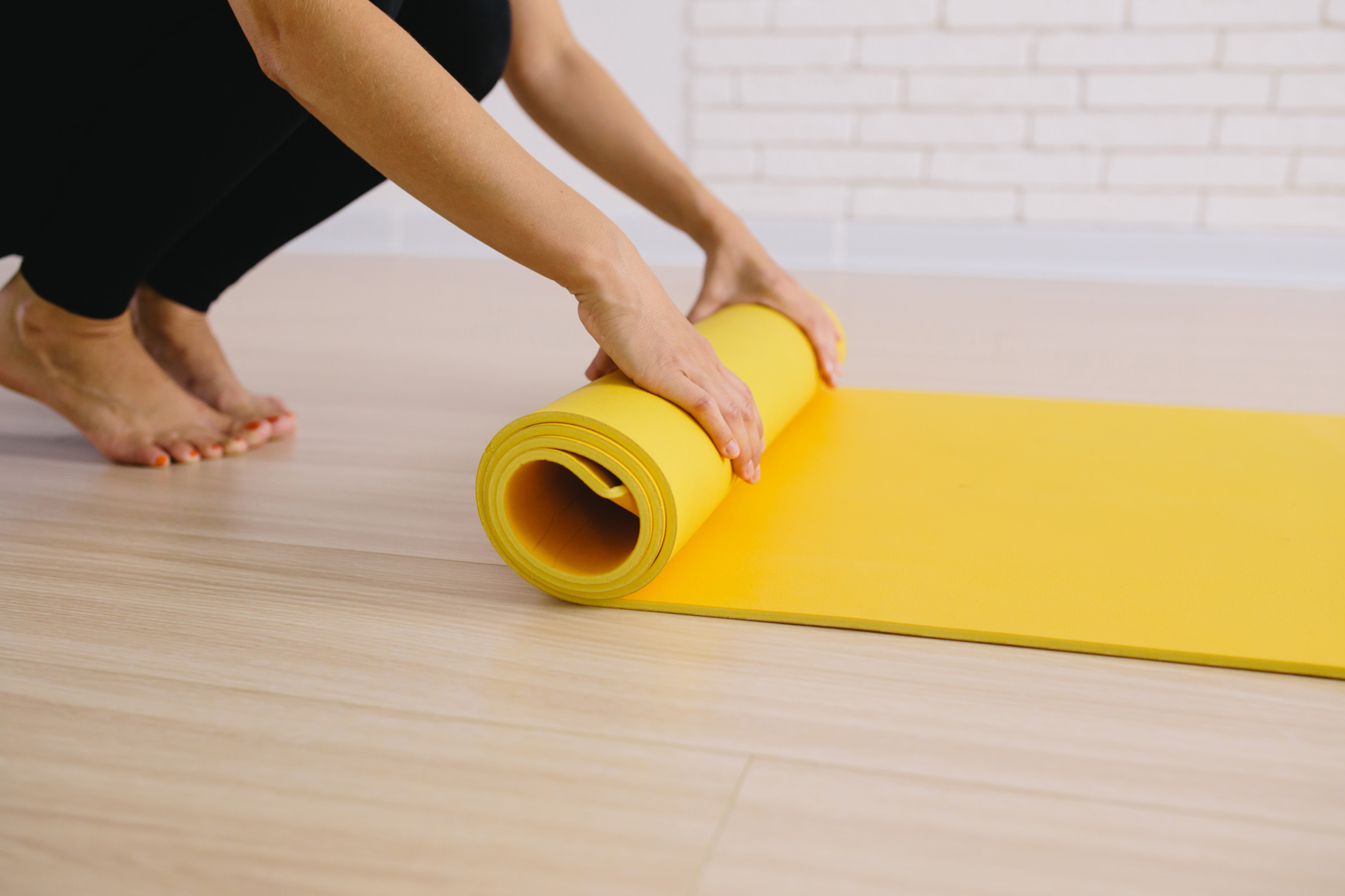 Eine Person rollt eine gelbe Yogamatte auf einem hellen Holzboden aus. Im Hintergrund ist eine helle Wand mit Fliesen zu sehen.
