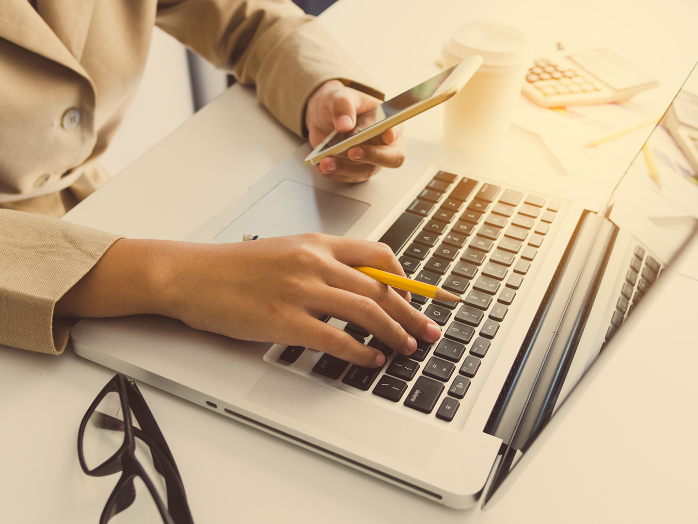 Eine Hand tippt auf einer Laptop-Tastatur, während die andere ein Smartphone hält. Im Hintergrund sind eine Tasse Kaffee und ein Taschenrechner sichtbar. Die Szene vermittelt eine Arbeitsumgebung.