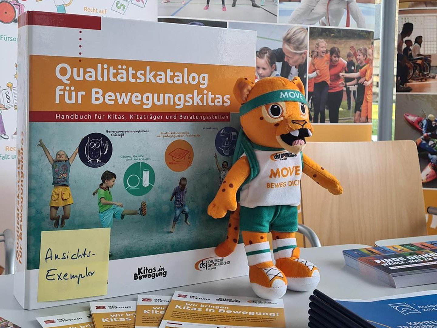 Ein großer Katalog mit dem Titel "Qualitätskatalog für Bewegungskitas" liegt auf einem Tisch. Daneben steht ein Plüschmaskottchen in Sportkleidung. Auf einem Zettel steht "Ansichts-Exemplar". Im Hintergrund sind weitere Materialien und Bilder zu sehen.