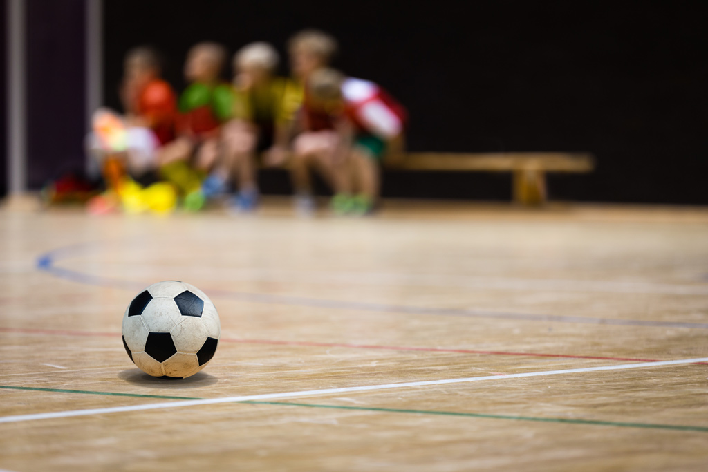 Ein Fußball liegt auf dem Boden einer Sporthalle. Im Hintergrund sind unscharf mehrere Kinder in bunten Trikots zu sehen, die auf einer Bank sitzen.