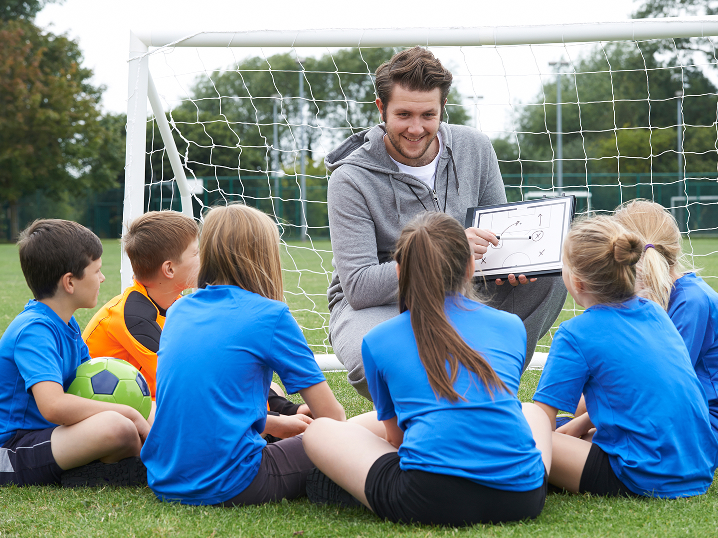 Ein Trainer erklärt einer Gruppe von Kindern auf einem Fußballfeld eine Spielstrategie. Die Kinder, in blauen T-Shirts, sitzen im Halbkreis vor dem Trainer, der ein Taktikboard hält. Im Hintergrund ist ein Fußballtor sichtbar.