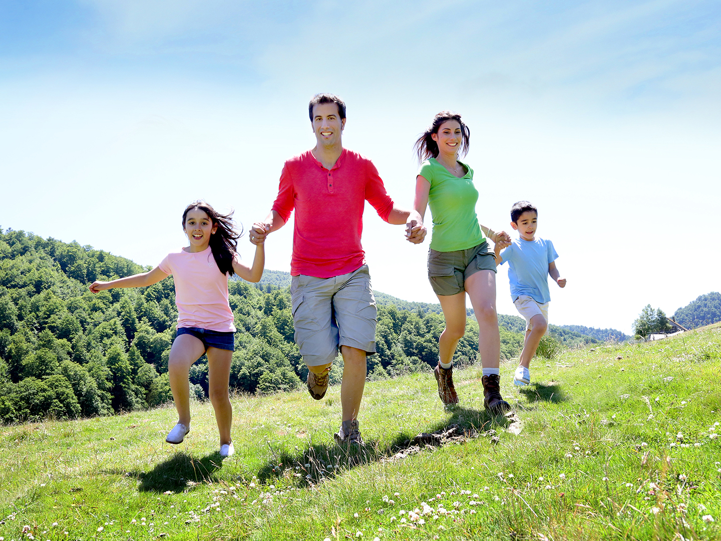 Eine Familie mit zwei Erwachsenen und zwei Kindern läuft fröhlich über eine grüne Wiese. Die Sonne scheint, und im Hintergrund sind sanfte Hügel und Bäume zu sehen. Alle tragen lässige Kleidung und haben Spaß beim Spielen im Freien.