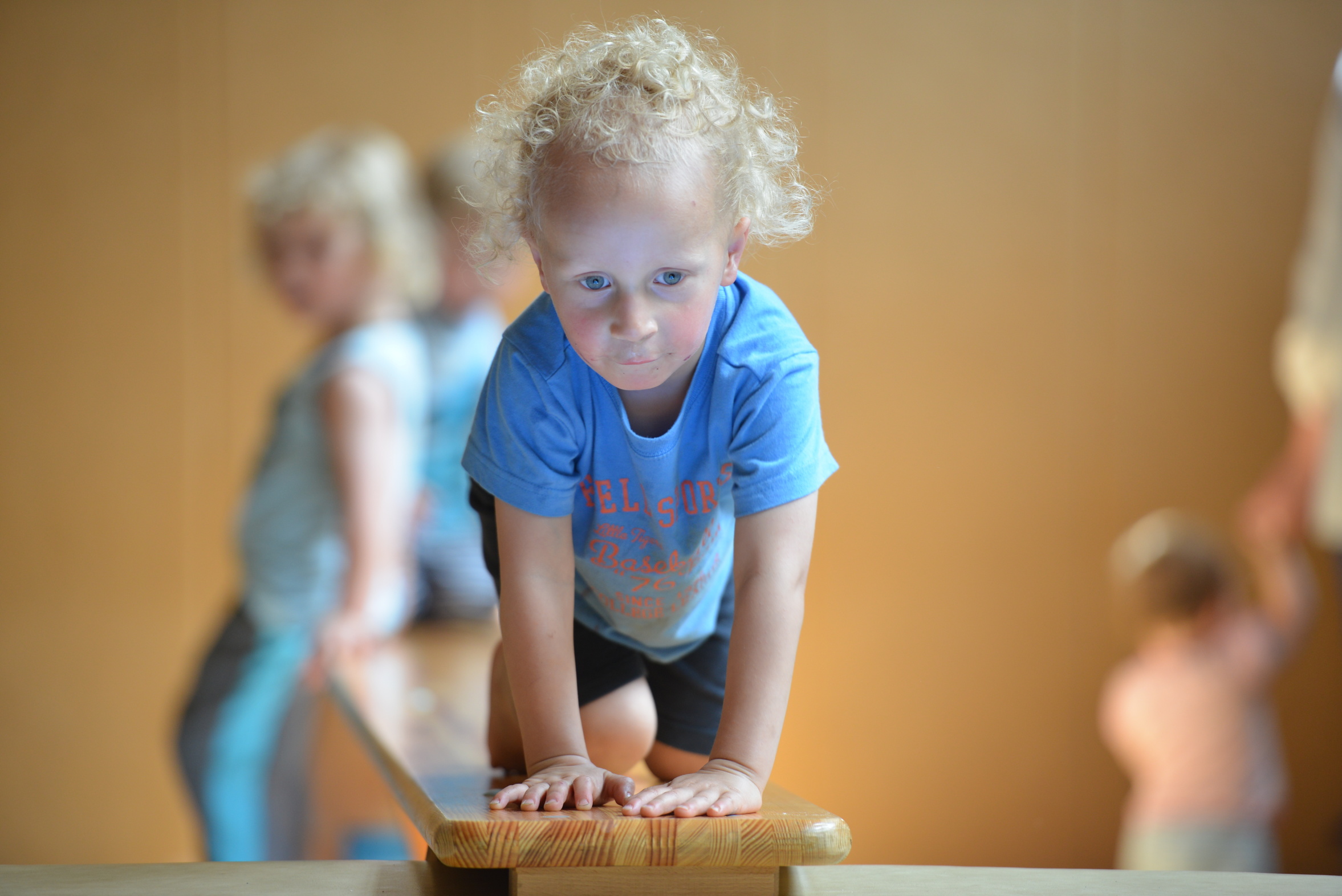 Ein kleiner Junge mit lockigem, blondem Haar krabbelt auf einem Holzbrett. Er trägt ein blaues T-Shirt und schaut konzentriert. Im Hintergrund sind weitere Kinder zu sehen, die ebenfalls spielen. Die Umgebung wirkt hell und freundlich.