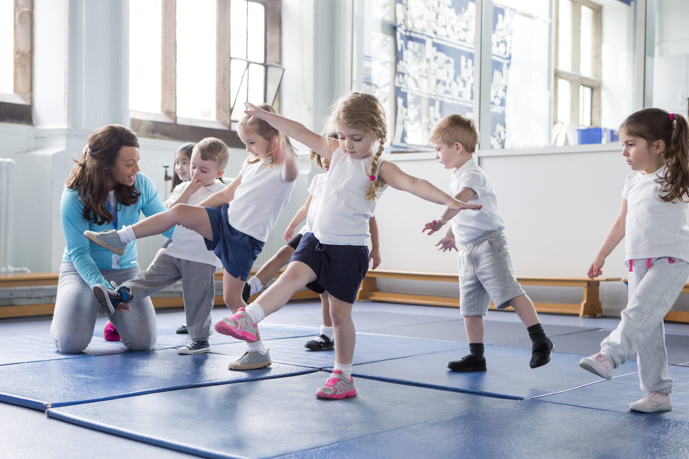 Eine Gruppe von Kindern in Sportkleidung übt Bewegungen auf einer blauen Turnmatte. Eine Erwachsene zeigt ihnen eine Übung, während die Kinder aufmerksam folgen. Die Szene vermittelt eine freundliche und aktive Lernumgebung.