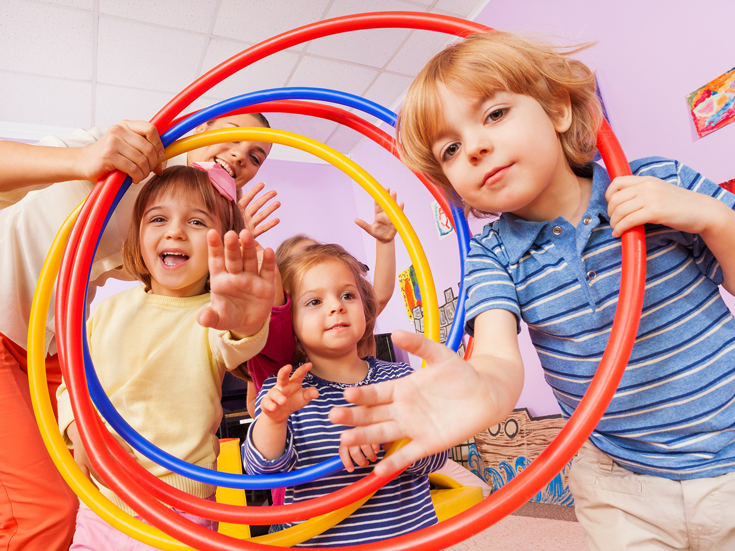 Eine Gruppe von Kindern spielt in einem Raum mit bunten Hula-Hoops. Zwei Kinder lächeln und strecken die Hände aus, während ein weiteres Kind hinter ihnen steht. Die Atmosphäre ist fröhlich und spielerisch.