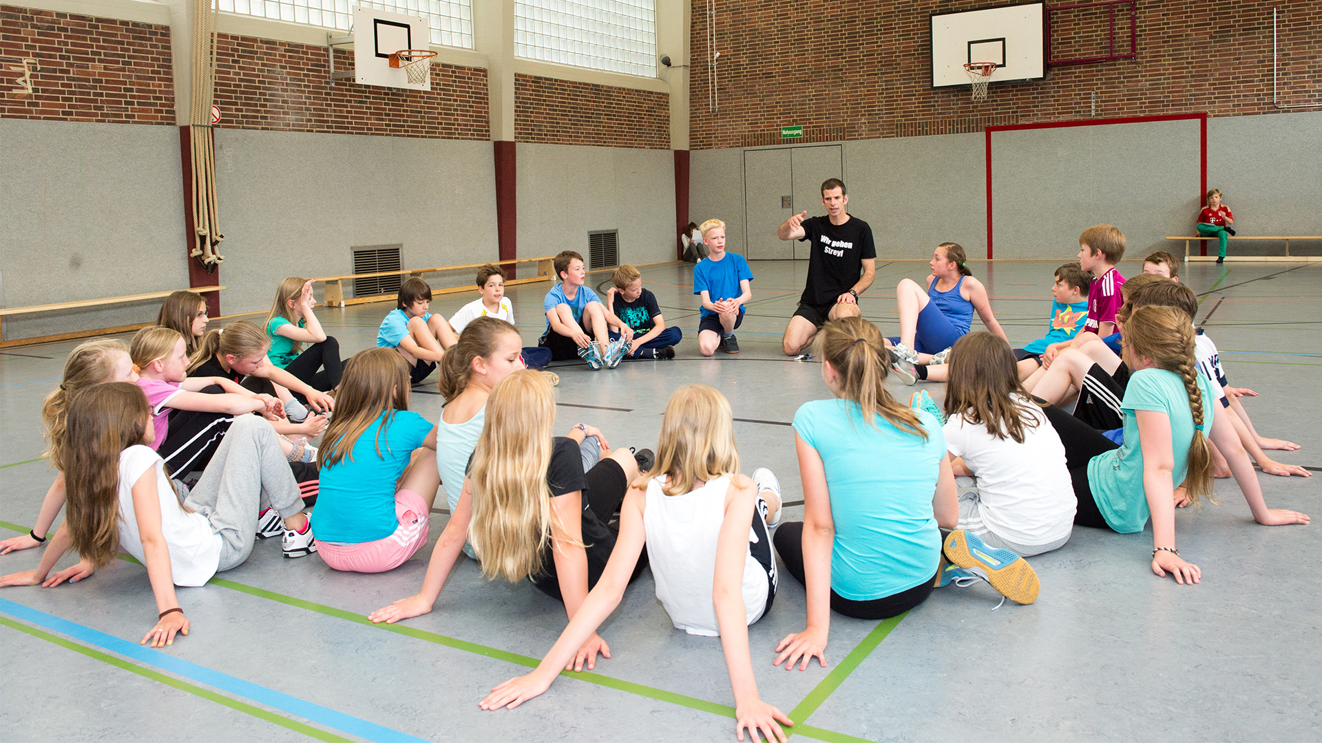 Kinder sitzen im Kreis in einer Turnhalle, eine Person in Sportkleidung spricht zu ihnen, Aktivität im Rahmen von 'Bewegt den ganzen Tag'