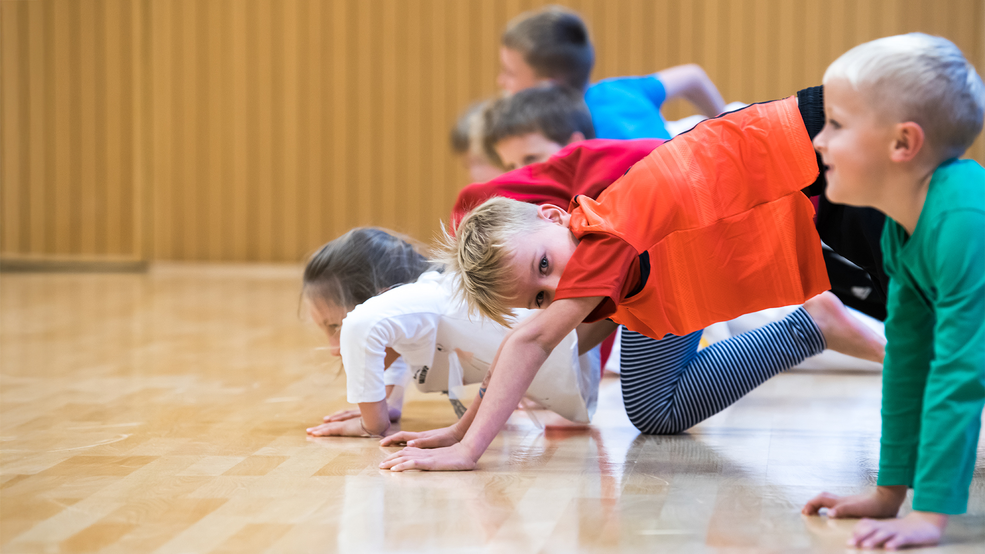 Kinder machen Liegestütze in einer Sporthalle mit Holzvertäfelung an der Wand