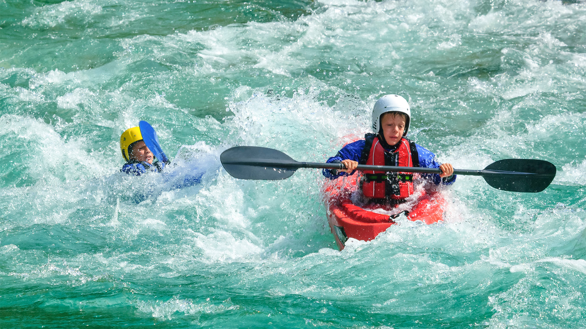 Zwei Kinder beim Wildwasser-Kajakfahren, eine im roten Kajak mit Helm und Schwimmweste, die andere schwimmt mit Helm im Wasser.