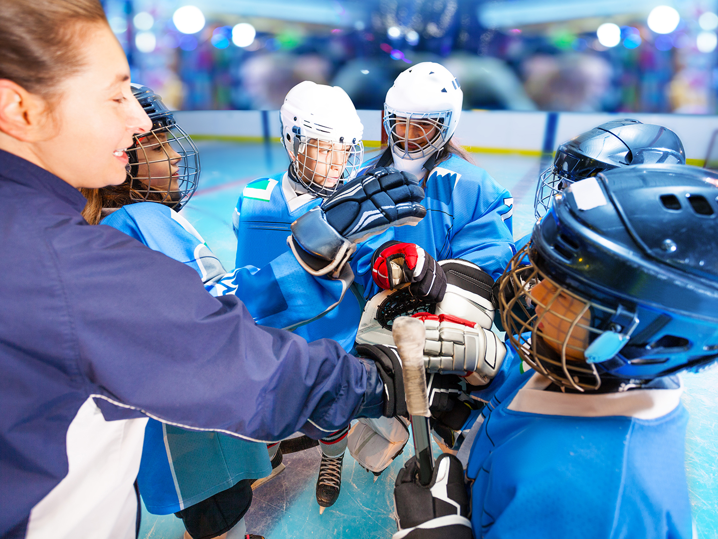 Eine Trainerin steht mit einer Gruppe von vier Eishockeyspielern auf dem Eis. Alle tragen Schutzausrüstung und blaue Trikots. Die Trainerin gibt Anweisungen, während die Spieler aufmerksam zuhören und sich auf das Spiel vorbereiten. Im Hintergrund sind unscharfe Lichter und eine Eishockeyarena zu sehen.