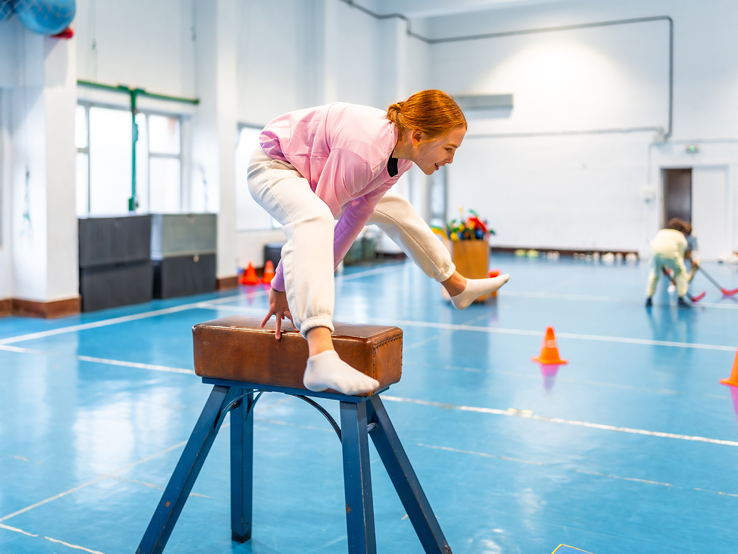 Eine junge Person mit roten Haaren springt über einen Turnbock in einer Sporthalle. Der Boden ist blau und es sind einige orangefarbene Pylonen im Hintergrund zu sehen. Ein weiterer Teilnehmer ist im Hintergrund mit einem Sportgerät beschäftigt.
