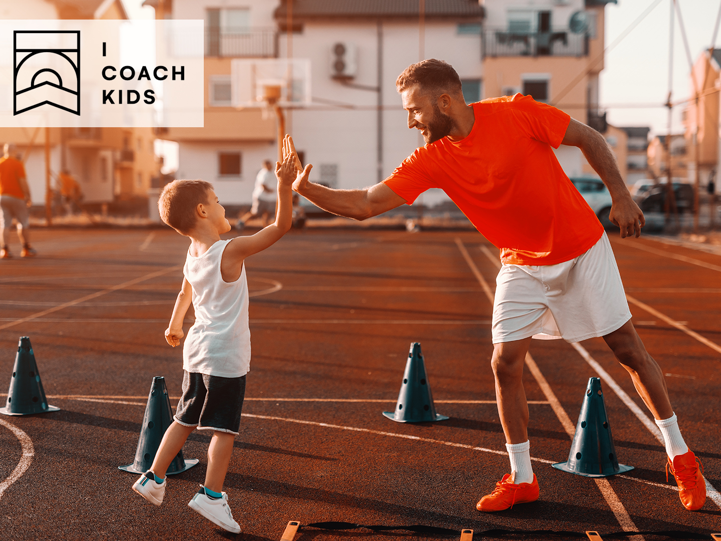 Ein Trainer und ein Junge geben sich auf einem Sportplatz ein High-Five. Der Trainer trägt ein rotes T-Shirt und Shorts, der Junge ein weißes Tanktop und schwarze Shorts. Um sie herum stehen blaue Hütchen. Im Hintergrund sind Basketballkörbe und Gebäude sichtbar.
