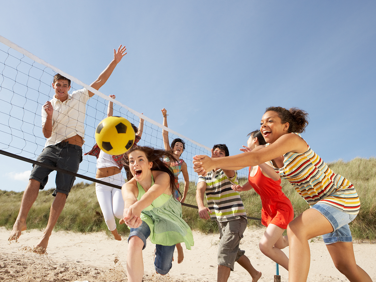 Eine Gruppe von jungen Menschen spielt Beachvolleyball am Strand. Einige springen, um den Ball zu erreichen, während andere jubeln. Die Atmosphäre ist fröhlich und lebhaft, mit blauem Himmel und Sand im Hintergrund.