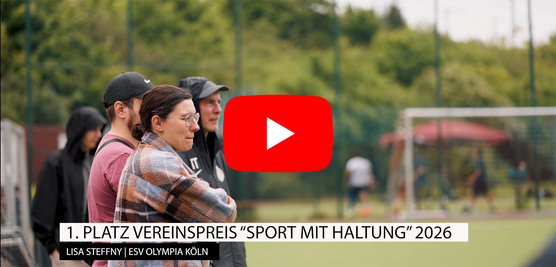 Mehrere Personen stehen am Rand eines Fußballplatzes und schauen auf das Spielfeld; im Hintergrund sind ein Tor und weitere Spielerinnen und Spieler zu sehen. Über dem Bild eingeblendet: „1. Platz Vereinspreis ‚Sport mit Haltung‘ 2026 – Lisa Steffny | ESV Olympia Köln“