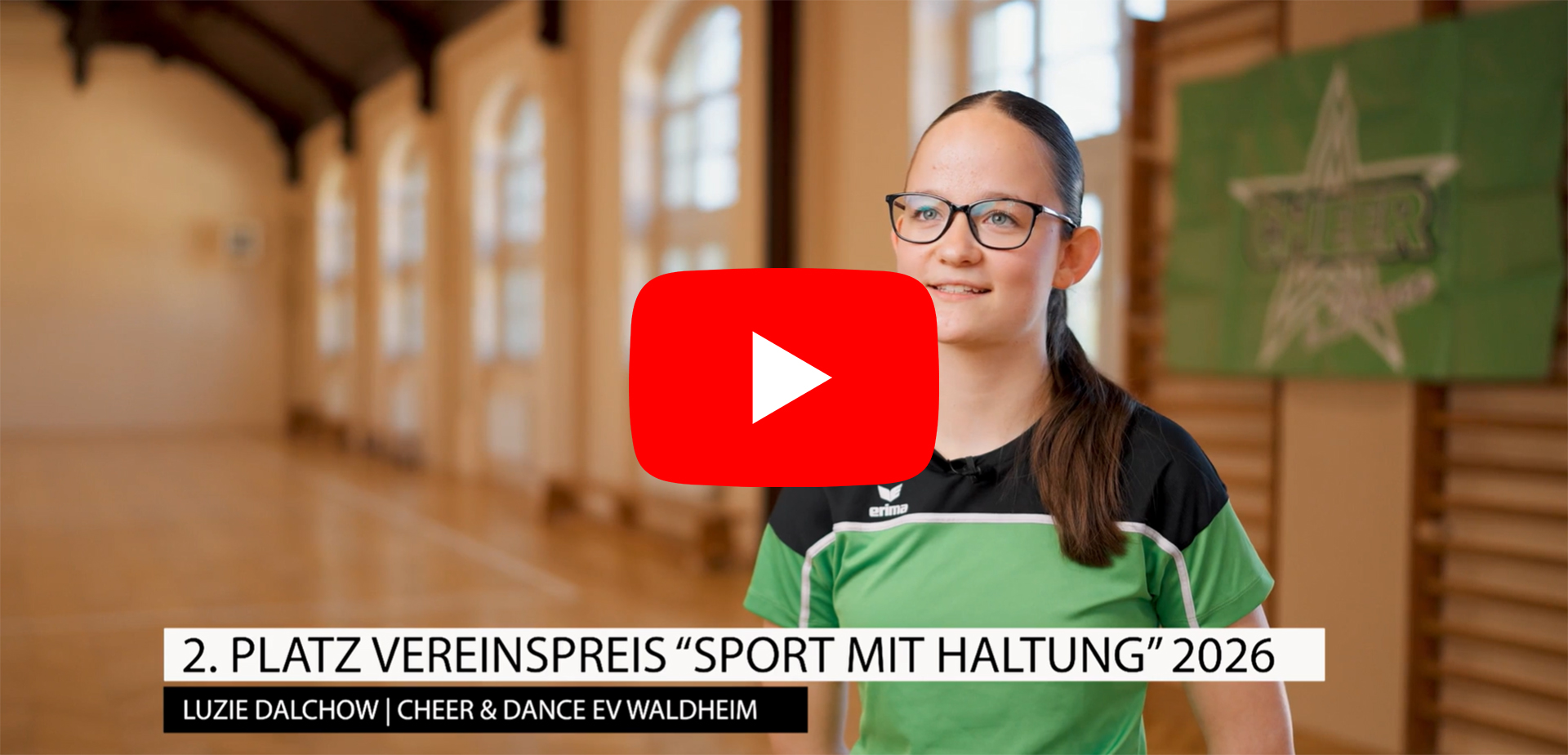 Eine Person in Sportkleidung steht in einer Turnhalle mit hohen Fenstern und Holzsprossenwand im Hintergrund. Rechts ist ein grünes Plakat mit Sternmotiv zu sehen. Im Bild eingeblendet: „2. Platz Vereinspreis ‚Sport mit Haltung‘ 2026 – Luzie Dalchow | Cheer & Dance e.V. Waldheim“.