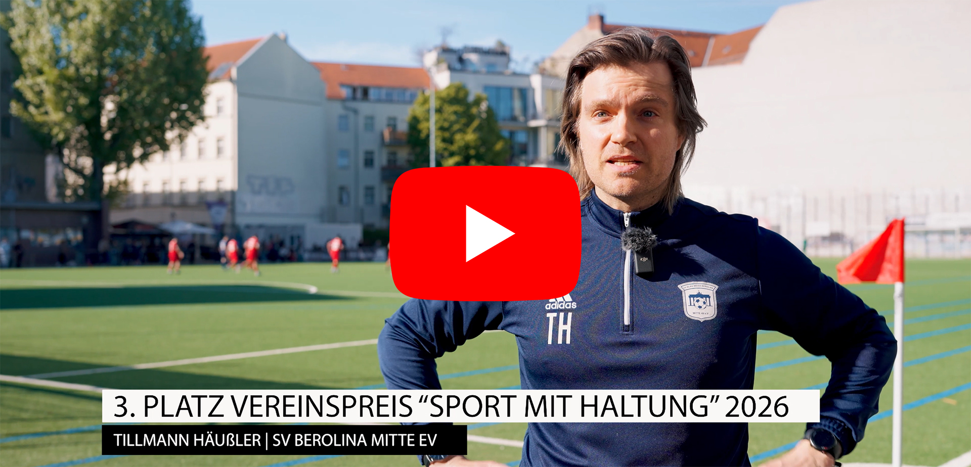 Eine Person in Sportkleidung steht auf einem Fußballplatz im Vordergrund; im Hintergrund sind weitere Personen beim Training sowie Gebäude am Rand des Platzes zu sehen. Im Bild eingeblendet: „3. Platz Vereinspreis ‚Sport mit Haltung‘ 2026 – Tillmann Häußler | SV Berolina Mitte e.V.“.
