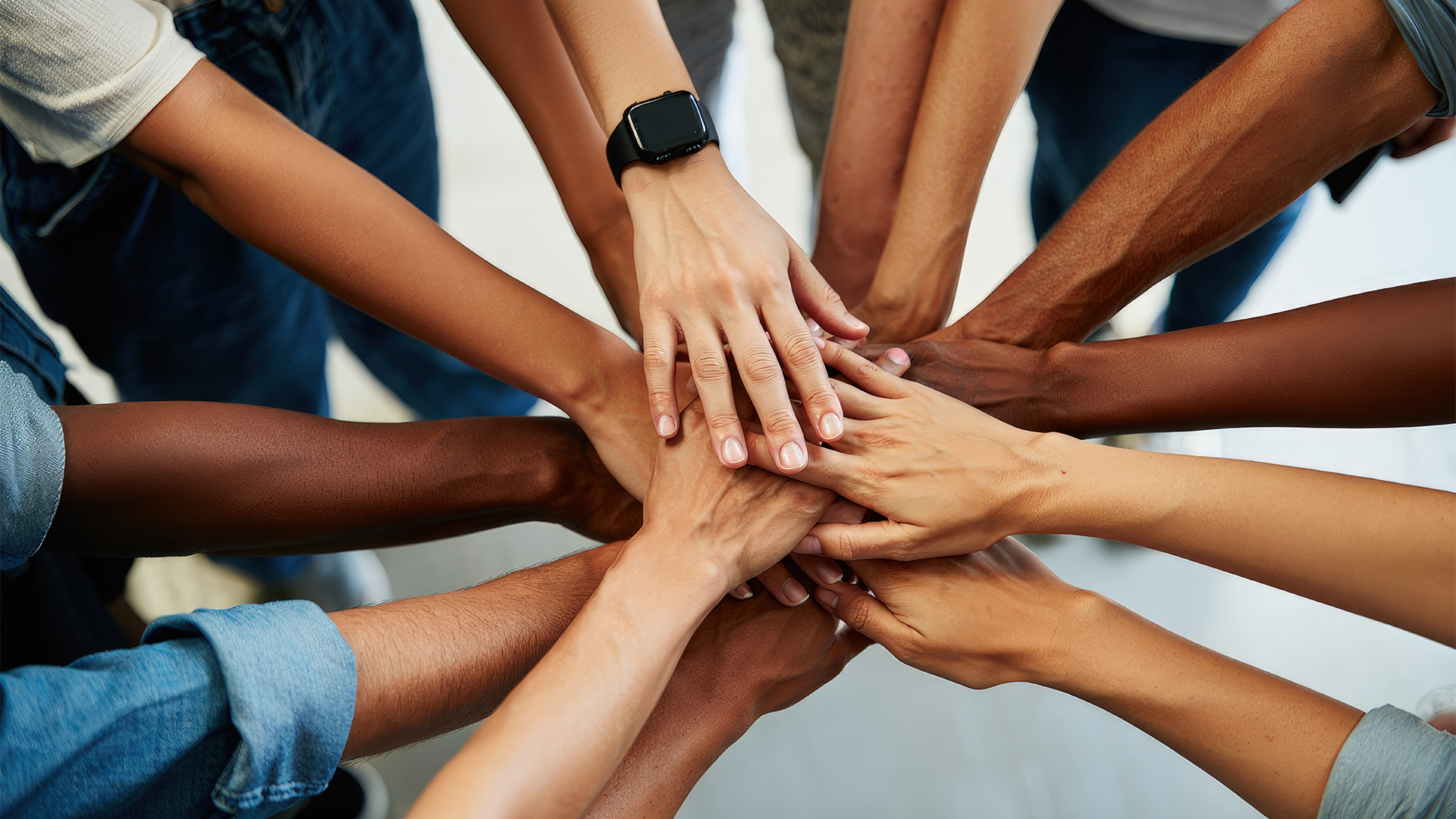 Mehrere Hände unterschiedlicher Hauttöne liegen übereinander in der Mitte eines Kreises. Die Hände sind in verschiedenen Positionen angeordnet, was Gemeinschaft und Zusammenarbeit symbolisiert.