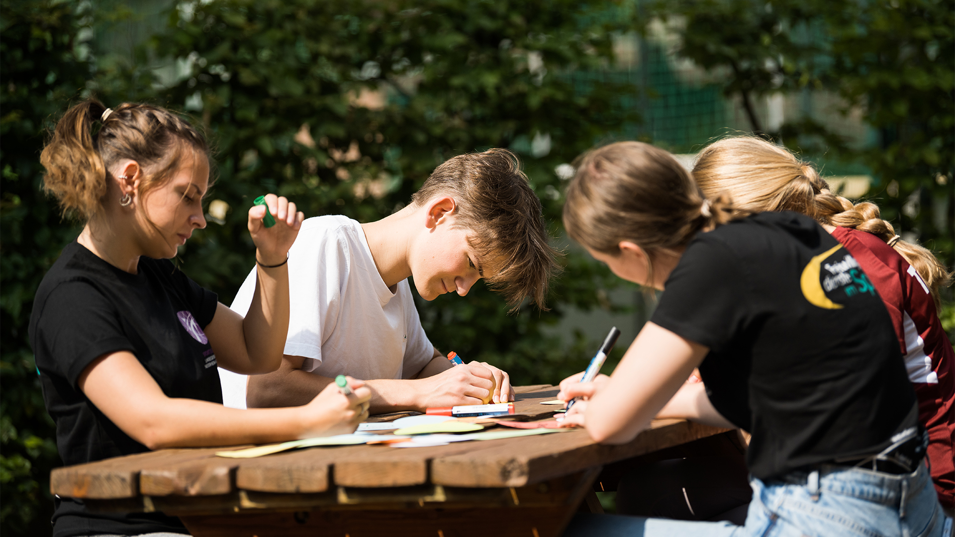 Vier junge Menschen sitzen an einem Tisch im Freien und zeichnen. Sie verwenden verschiedene Stifte und Papier. Die Sonne scheint, und im Hintergrund sind grüne Sträucher zu sehen.