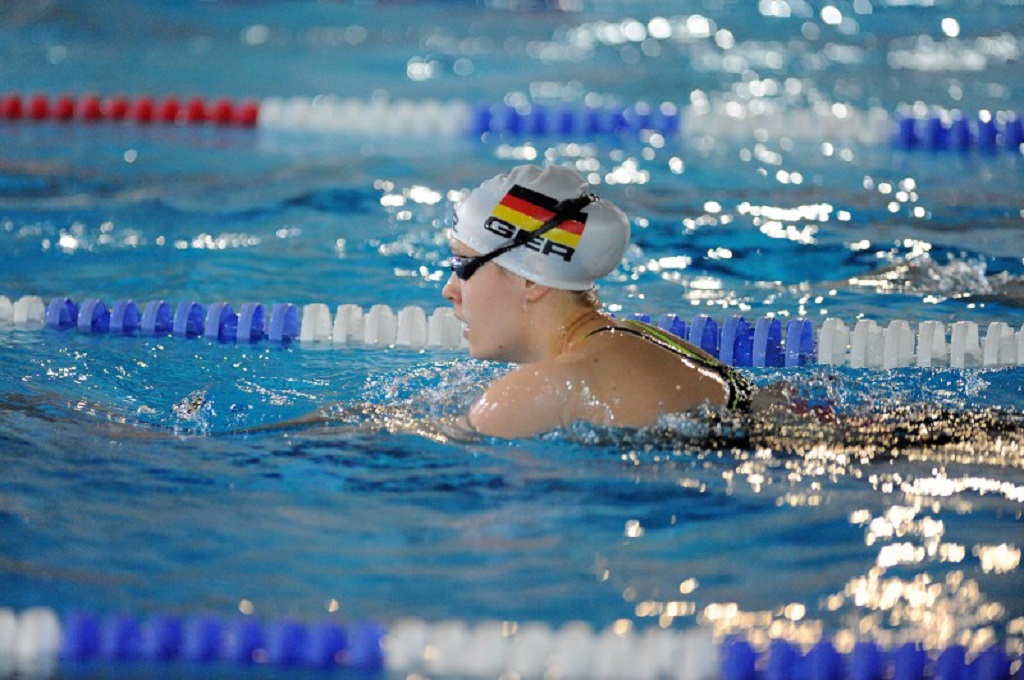 Eine Schwimmerin mit einer Badekappe in den Farben der deutschen Flagge schwimmt im Wettkampfbecken. Das Wasser spritzt um sie herum, während sie sich auf ihre Technik konzentriert.