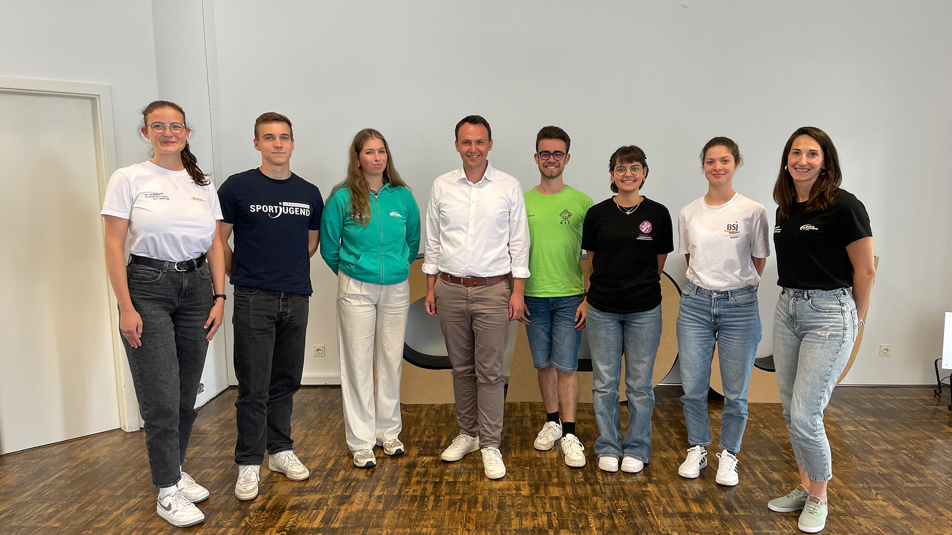 Gruppe von acht Personen steht in einem Raum mit Holzboden, darunter der CDU-Bundestagsabgeordnete Pascal Reddig in weißem Hemd und beiger Hose, umgeben von jungen Erwachsenen in Freizeitkleidung.
