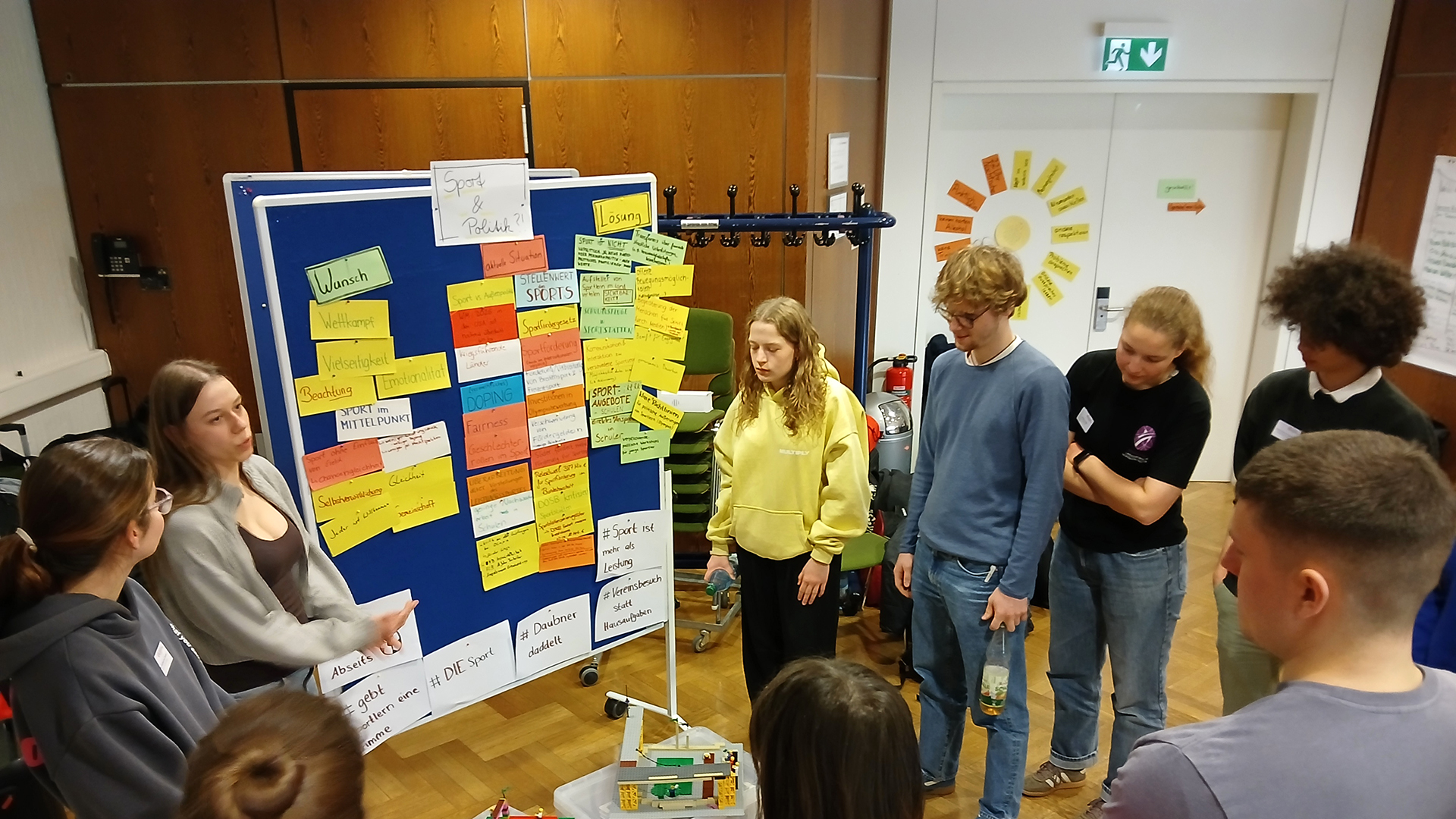 Gruppe junger Menschen steht in einem Workshopraum um eine Pinnwand mit Karten und Notizen und diskutiert gemeinsam Ideen zur aktiven Mitgestaltung im Sport.