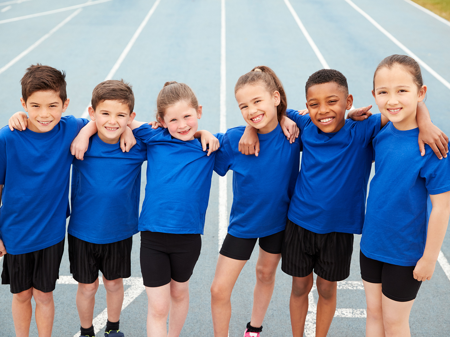 Sechs Kinder in Sportkleidung stehen auf einer Laufbahn und legen sich gegenseitig die Arme über die Schultern.