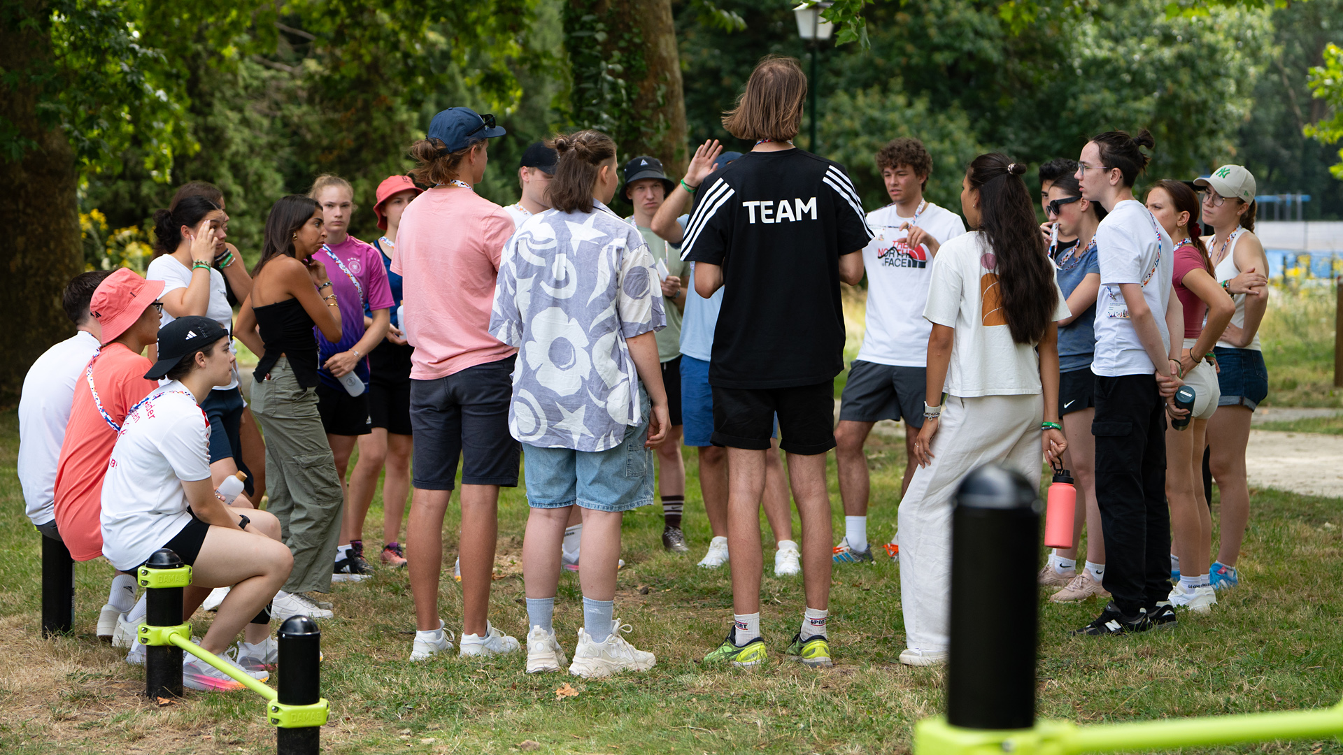 Eine Gruppe von Jugendlichen steht im Freien zusammen, einige sitzen auf dem Boden. Eine Person mit einem T-Shirt mit der Aufschrift "TEAM" spricht zu den anderen. Die Umgebung ist grün und sonnig, mit Bäumen im Hintergrund.
