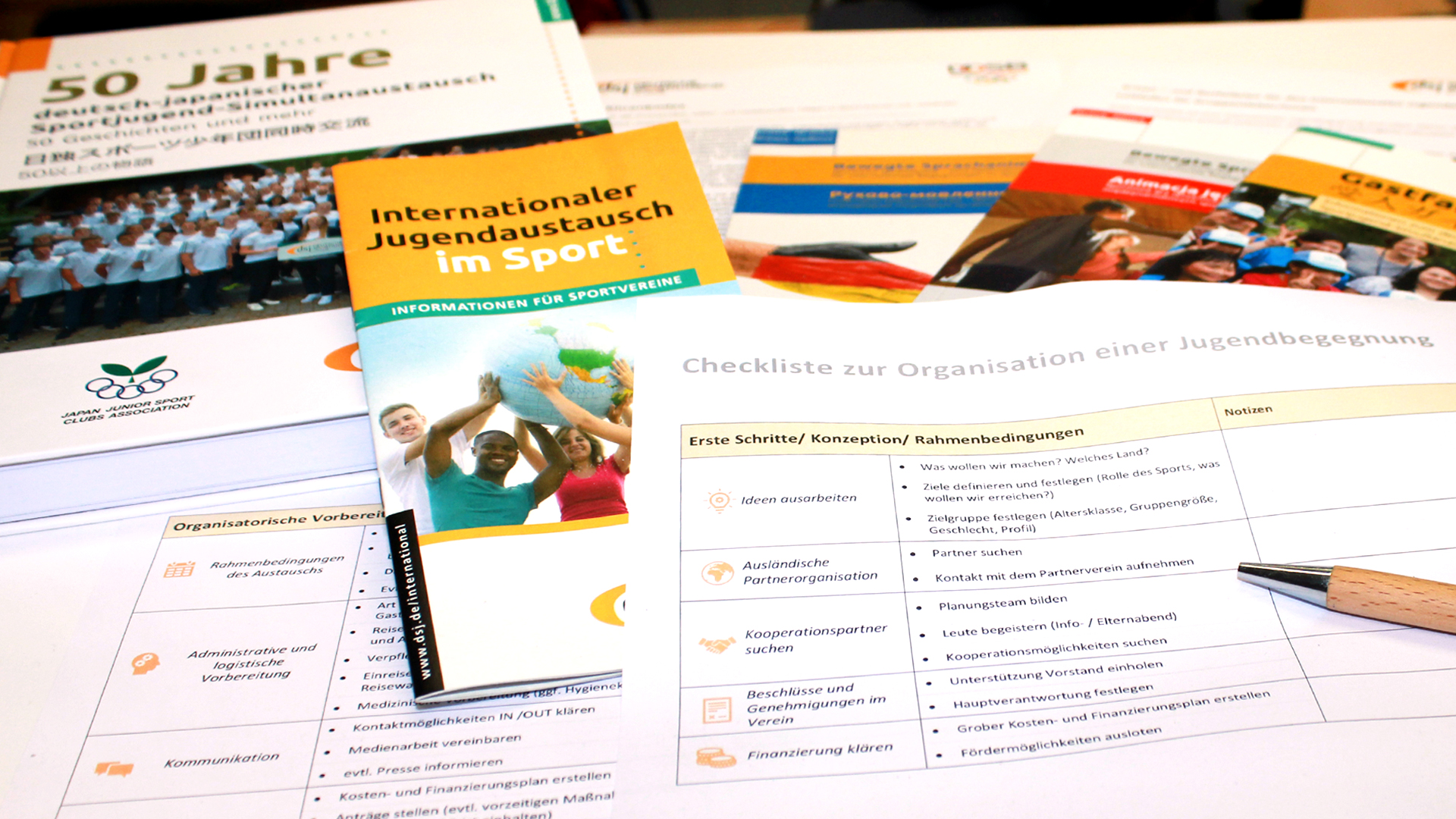 Auf einem Tisch liegen verschiedene Dokumente zum Thema internationaler Jugendaustausch im Sport. Darunter befindet sich eine Checkliste zur Organisation einer Jugendbegegnung, ein Informationsheft für Sponsoren und ein Jubiläumsheft über 50 Jahre. Ein Stift liegt neben den Unterlagen.