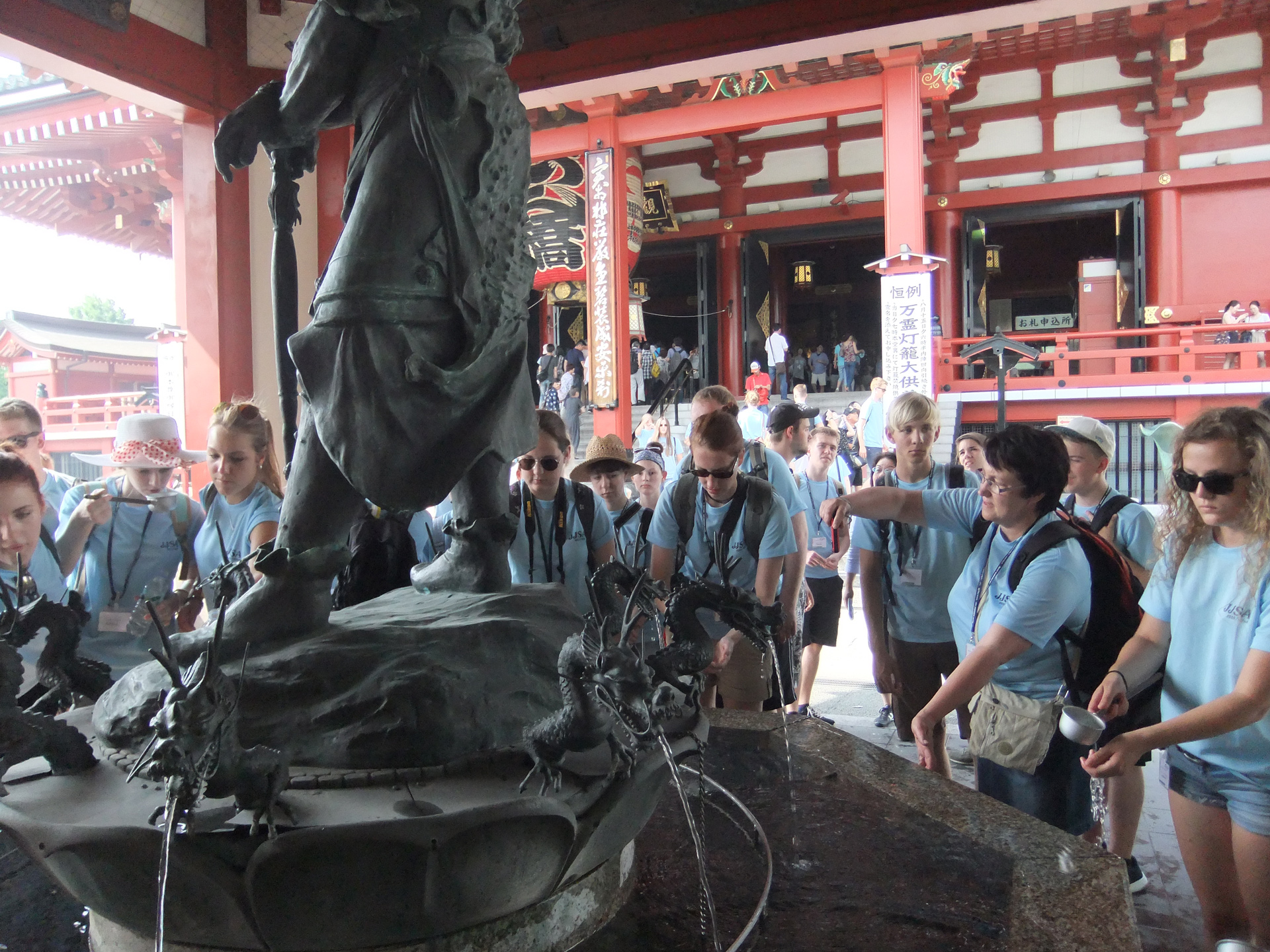 Eine Gruppe von Teilnehmenden in blauen T-Shirts steht um eine Statue in einem Tempel. Die Statue zeigt eine Figur, die von Wasserspeiern umgeben ist, aus denen Wasser sprudelt. Im Hintergrund sind weitere Tempelstrukturen sichtbar.