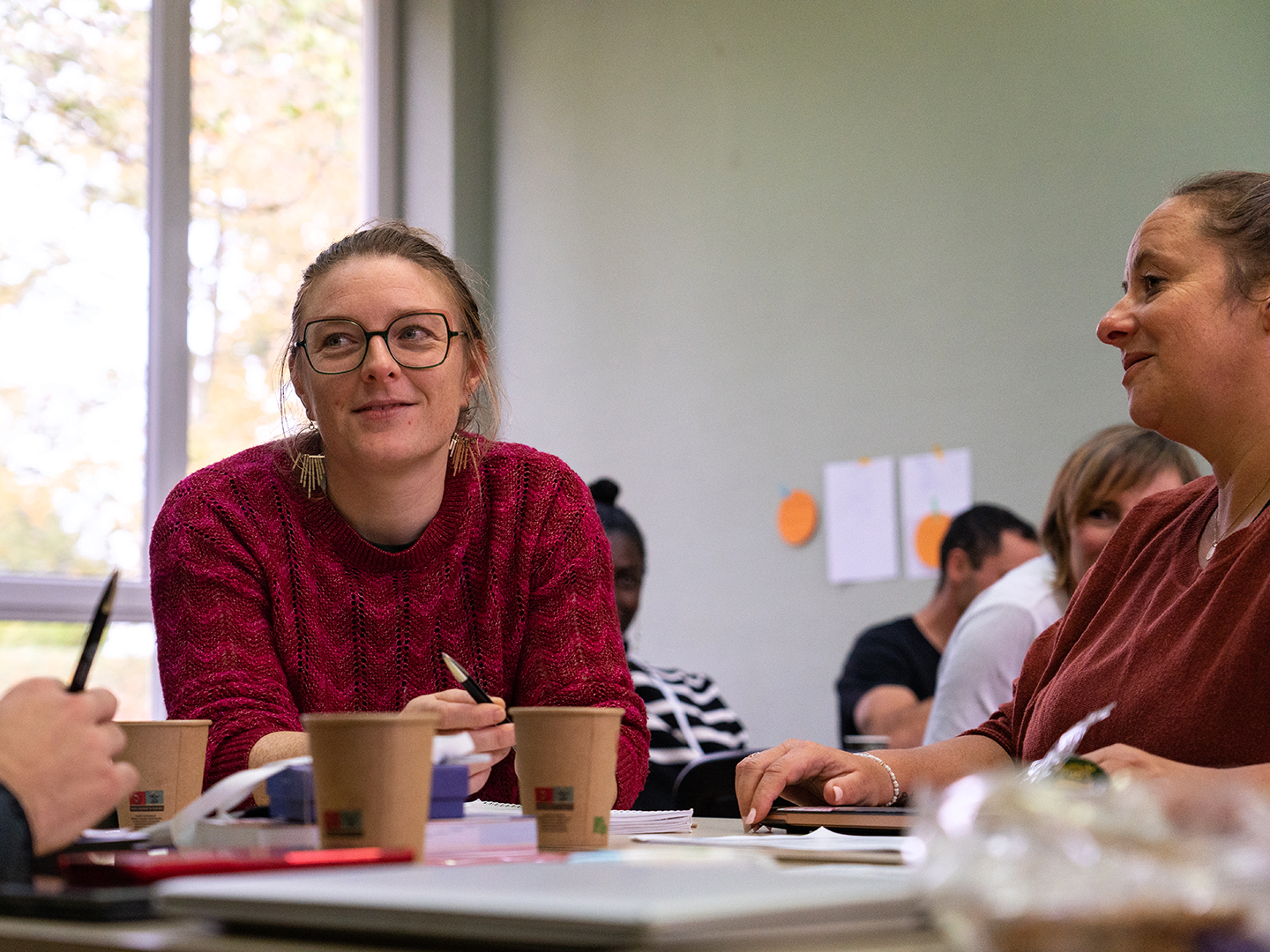 Eine Gruppe von Menschen sitzt an einem Tisch, diskutiert und arbeitet zusammen. Eine Frau mit Brille und rotem Pullover lächelt, während sie mit einer anderen Frau spricht. Auf dem Tisch stehen Kaffeebecher und Notizen. Im Hintergrund sind weitere Personen und einige Notizen an der Wand sichtbar.