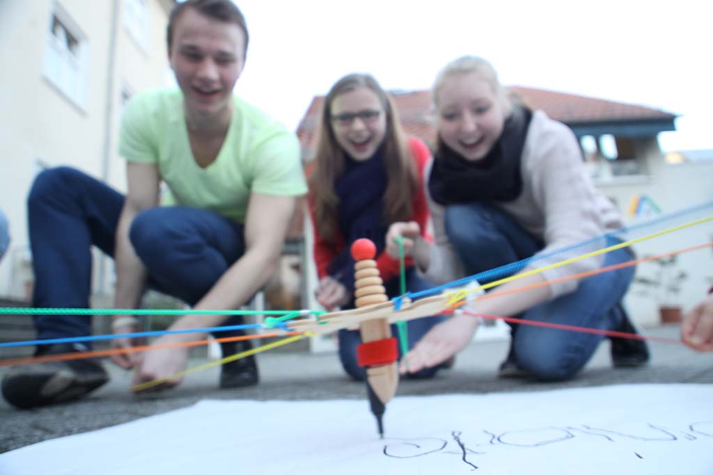 Drei Jugendliche sitzen auf dem Boden und lachen, während sie mit einem kreativen Zeichengerät arbeiten. Das Gerät besteht aus einem Bleistift, der von bunten Gummibändern gehalten wird, und es zeichnet auf ein großes Blatt Papier.