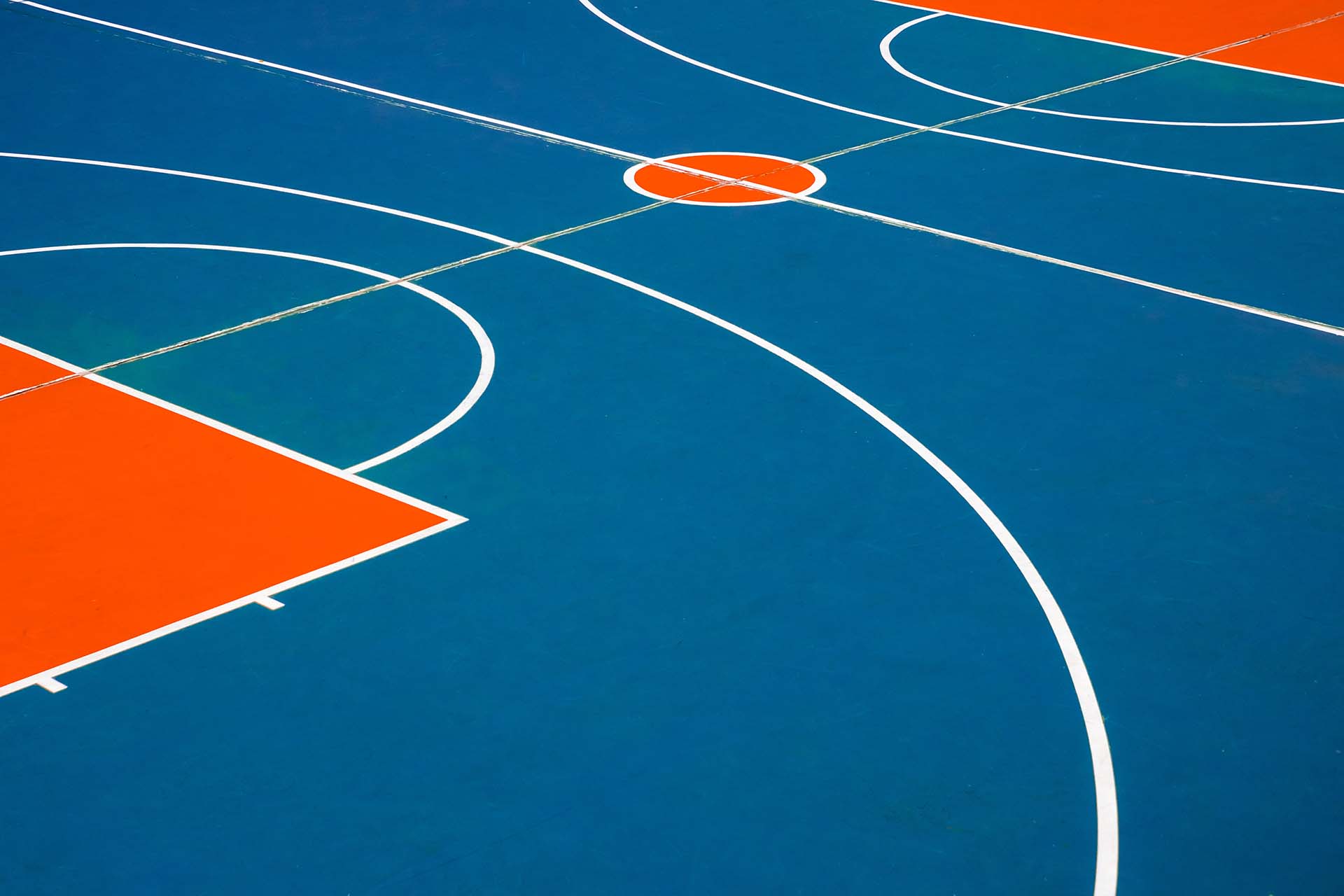 Ein Basketballplatz mit blauer Grundfarbe und orangefarbenen Bereichen. Die Linien sind weiß und zeigen die Spielfeldmarkierungen, einschließlich der drei-Punkte-Linie und des Mittelkreises.