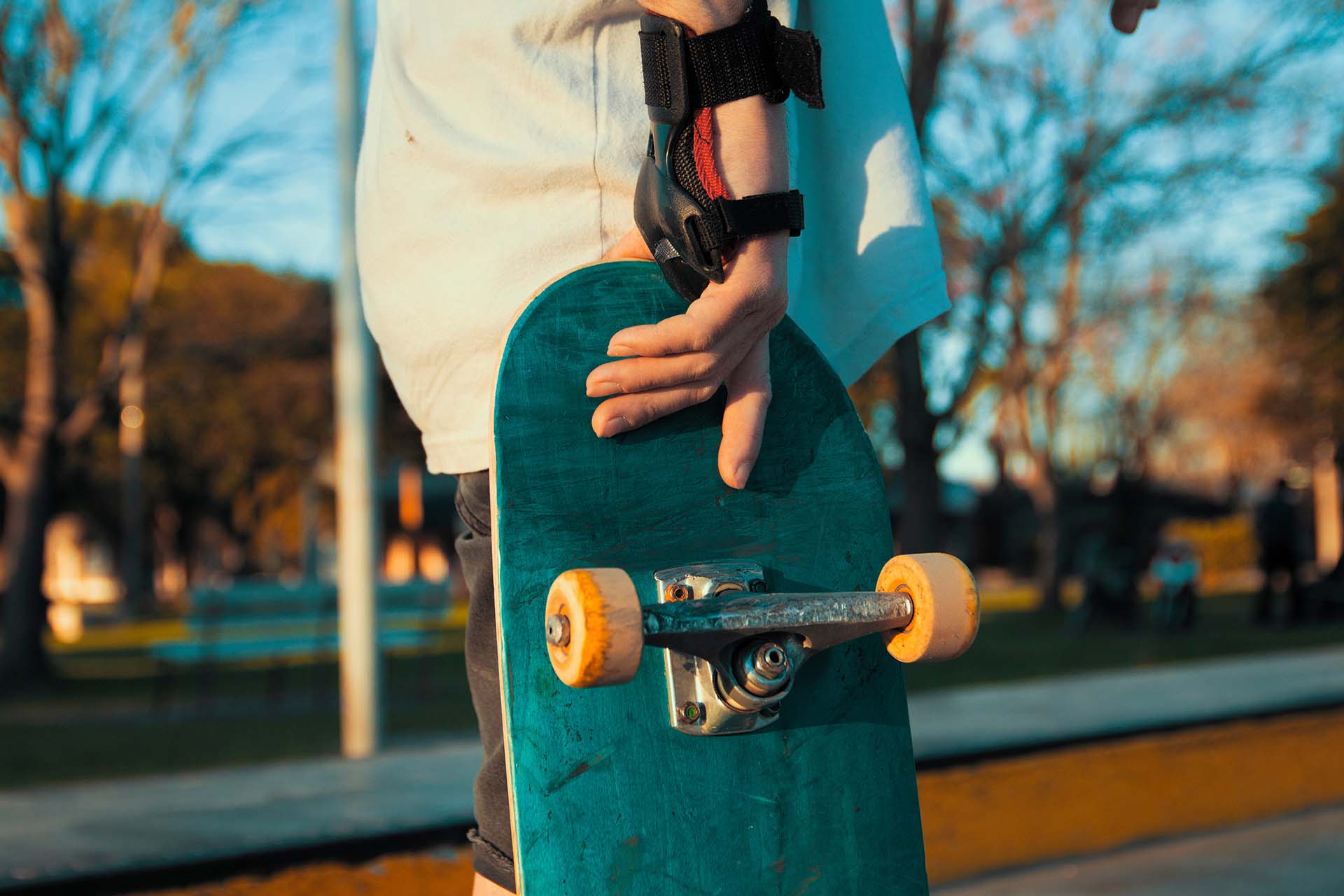 Ein Skateboarder hält ein Skateboard mit einer Hand. Das Board hat orangefarbene Rollen und eine grüne Oberfläche. Im Hintergrund sind Bäume und eine Parklandschaft sichtbar.