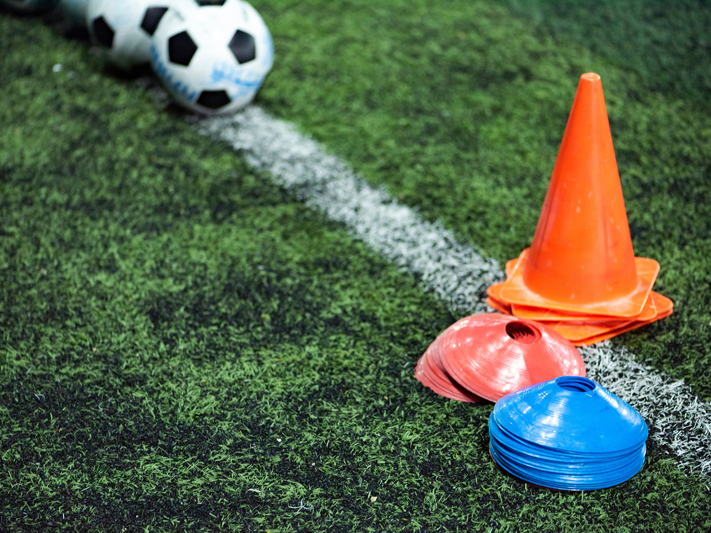 Auf einem Fußballfeld liegen mehrere Fußbälle neben einem orangefarbenen Kegel. Daneben befinden sich rote und blaue Markierungsscheiben auf dem grünen Kunstrasen.