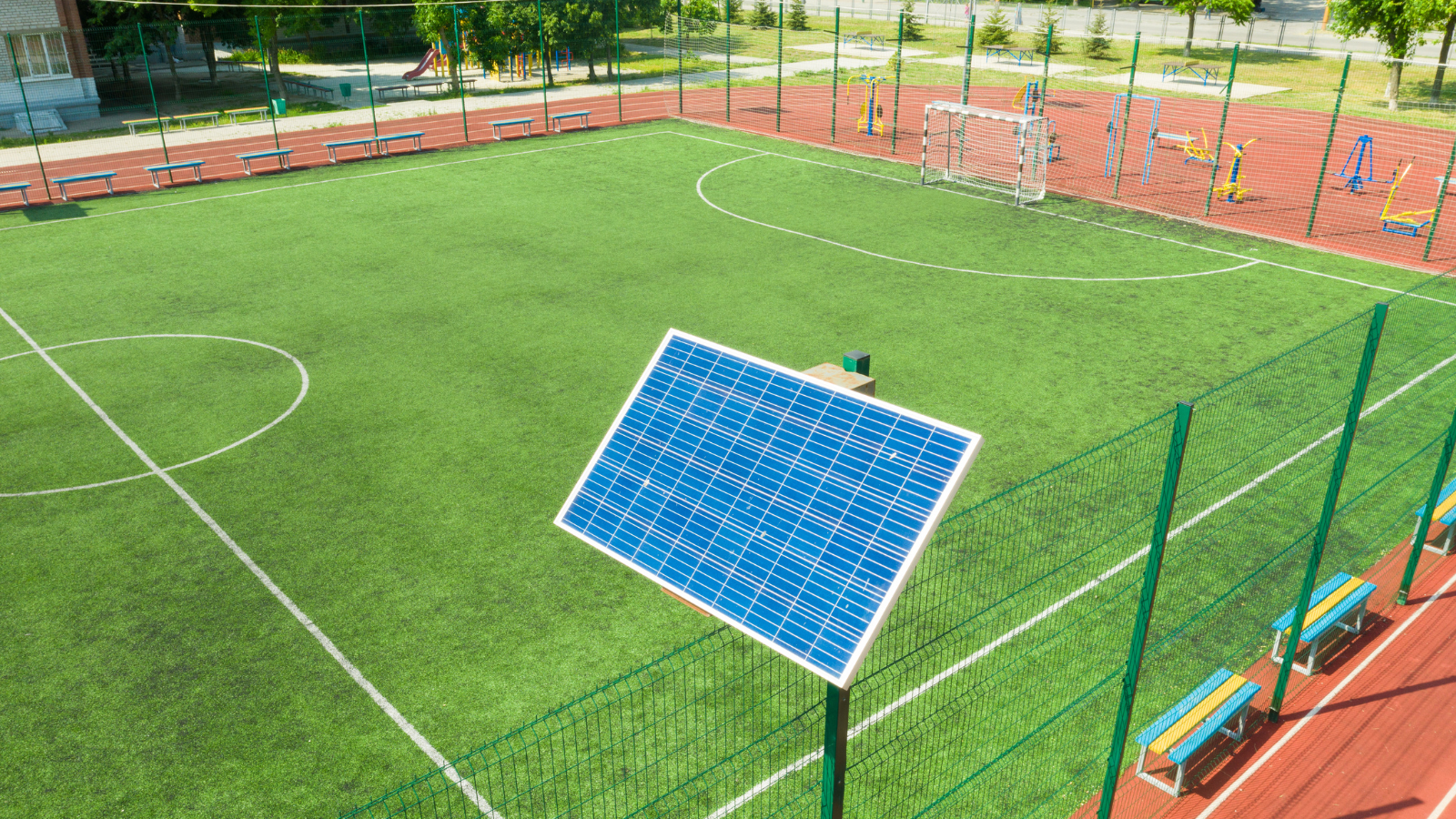 Ein Fußballplatz mit Kunstrasen, umgeben von einem Zaun. Im Vordergrund steht ein Solarpanel, das Energie erzeugt. Im Hintergrund sind Tore und Spielgeräte sichtbar.