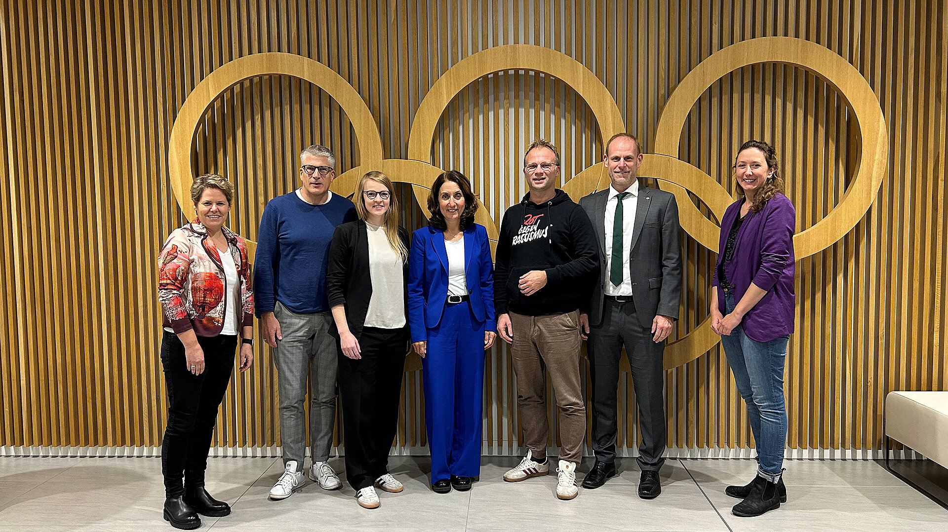 Eine Gruppe von sieben Personen steht vor einer Wand mit drei großen, goldfarbenen Ringen, die an die Olympischen Ringe erinnern. Die Wand besteht aus vertikal angeordneten, hellbraunen Holzlatten. Die Personen sind in einer Reihe aufgestellt, tragen unterschiedliche Kleidung: von einem blauen Anzug über einen schwarzen Pullover mit Aufdruck bis zu einem lila Oberteil. Einige tragen Schuhe in Weiß oder Schwarz. Die Szene wirkt formell und symbolisiert einen Dialog oder ein Treffen.
