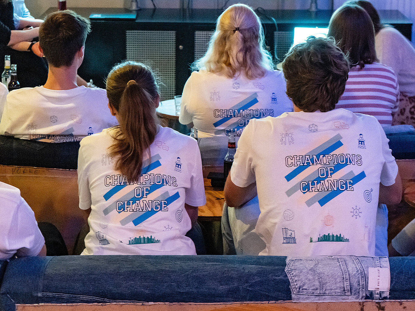 Eine Gruppe von Menschen sitzt auf einer Couch und trägt weiße T-Shirts mit der Aufschrift "CHAMPIONS OF CHANGE" auf dem Rücken. Die Personen sind von hinten zu sehen.