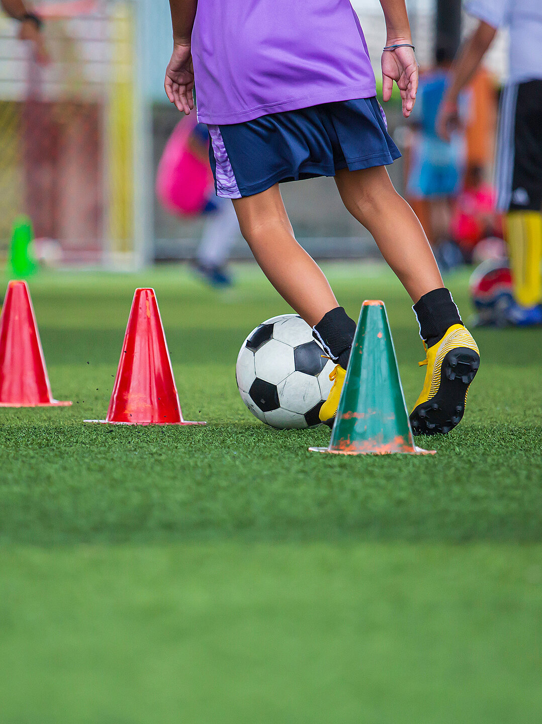 Ein Fußballspieler dribbelt einen Ball um aufgestellte Hütchen auf einem grünen Rasenplatz. Der Spieler trägt ein lila Trikot und schwarze Shorts. Im Hintergrund sind weitere Personen und Trainingsmaterialien zu sehen.